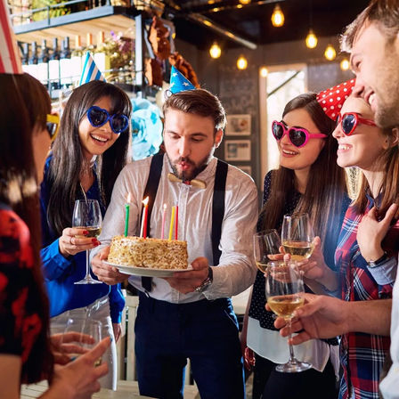Adults celebrating a birthday at an indoor bar — a man blowing out candles on a cake while friends in party hats and heart-shaped sunglasses cheer with wine glasses.