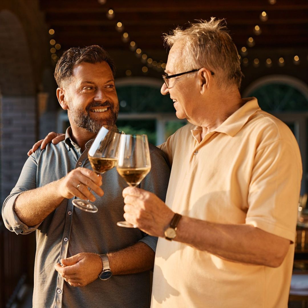 Two men—one older, one middle-aged—smiling and clinking glasses of white wine on a warm, string-lit outdoor patio at sunset.