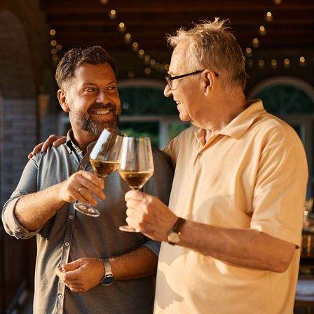 Two men—one older, one middle-aged—smiling and clinking glasses of white wine on a warm, string-lit outdoor patio at sunset.