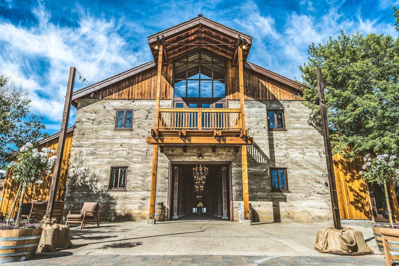 Sunlit rustic timber-and-concrete barn-style event venue with a wooden balcony, open central doorway revealing a cascading chandelier, string lights and potted planters under a bright blue sky.