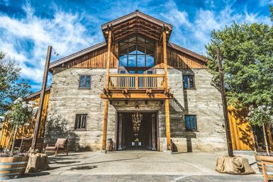 Sunlit rustic timber-and-concrete barn-style event venue with a wooden balcony, open central doorway revealing a cascading chandelier, string lights and potted planters under a bright blue sky.