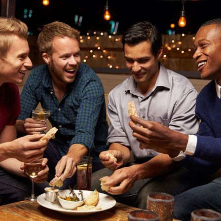 Four friends laughing and sharing bread and appetizers on an outdoor patio at night, enjoying cocktails under warm string lights