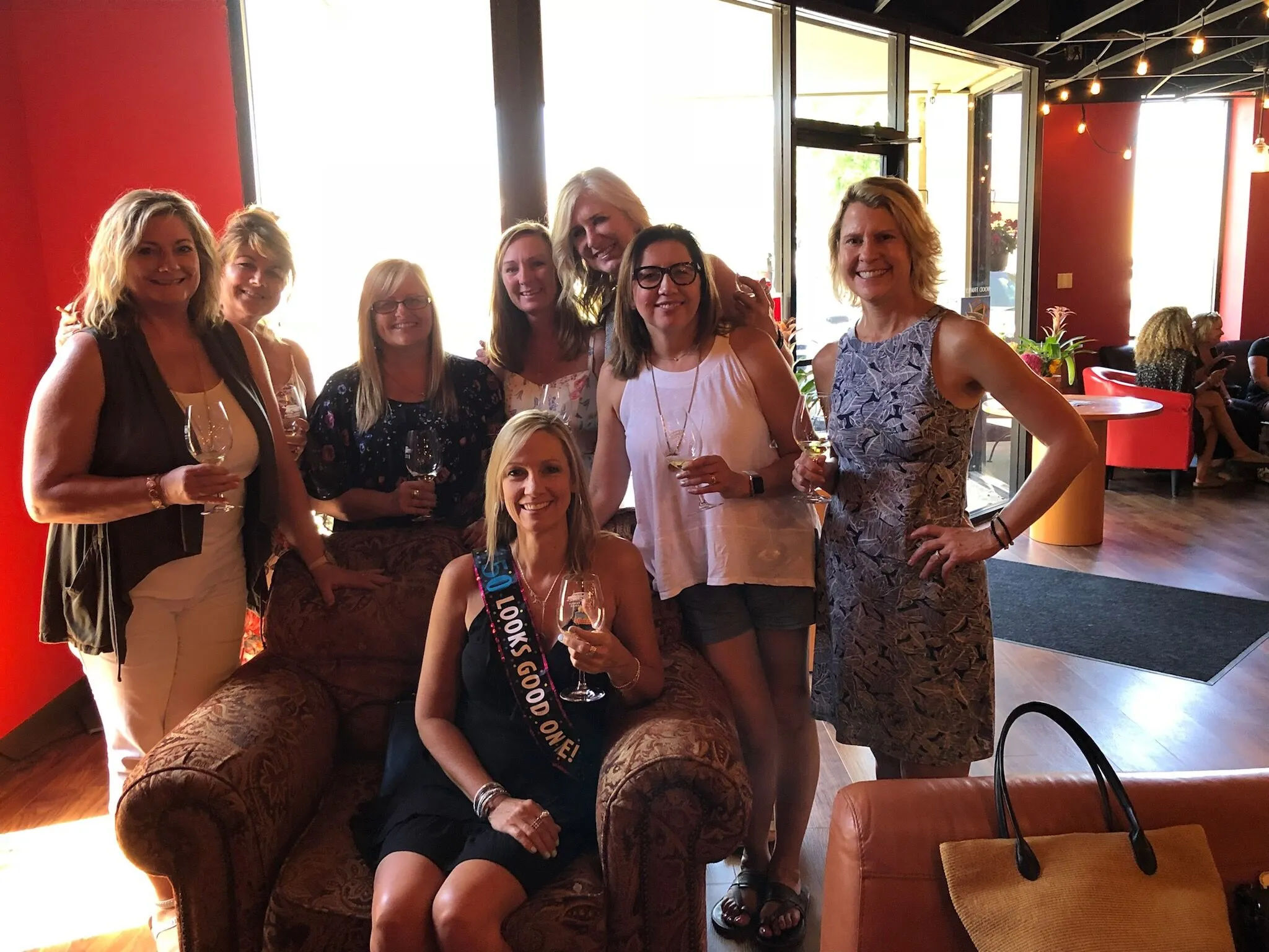 Cheerful group of eight women at a wine bar lounge holding wine glasses, one seated wearing a celebratory sash, warm red interior with cozy armchairs and string lights