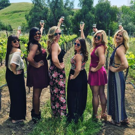 Six friends in sundresses and sunglasses standing among grapevines in a sunlit vineyard, smiling and raising wine glasses toward rolling green hills.