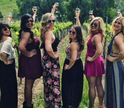 Six women in summer dresses and sunglasses raise wine glasses and pose along a sunny vineyard row, smiling and celebrating an outdoor wine-tasting.
