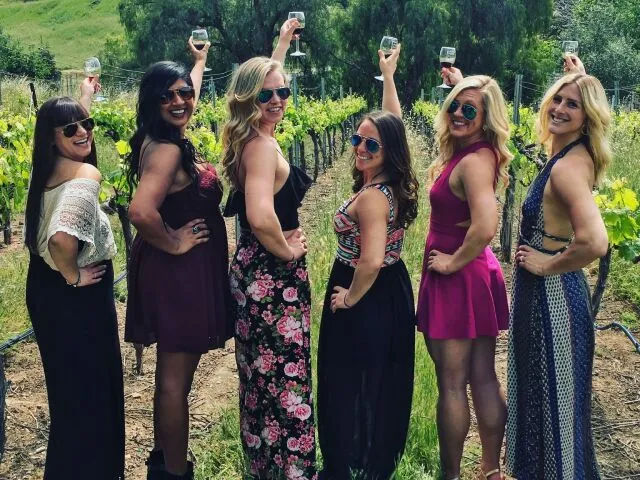 Six women in summer dresses and sunglasses raise wine glasses and pose along a sunny vineyard row, smiling and celebrating an outdoor wine-tasting.