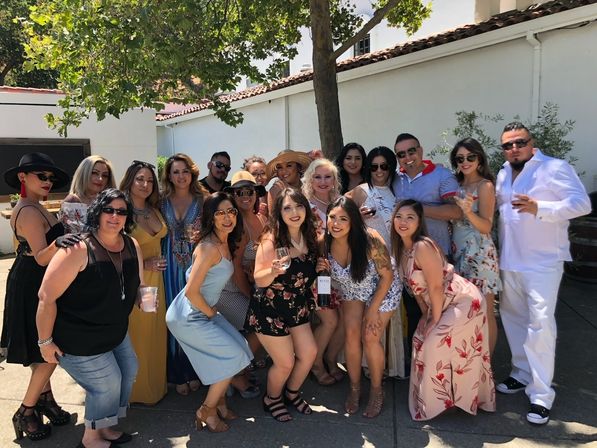 Group of adults posing on a sunny outdoor patio under a tree, wearing summer dresses and sunglasses, holding wine glasses and a bottle — casual daytime garden party.