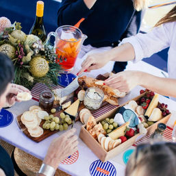 Al fresco charcuterie picnic with assorted cheeses, crackers, grapes and strawberries, jarred spreads and sliced bread, a pitcher of iced fruit tea and a bottle of white wine, hands reaching in around a floral centerpiece.