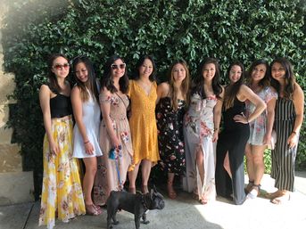 Nine women in colorful summer dresses posing for a group photo in front of an ivy-covered wall on a sunny patio, with a small black French bulldog at their feet.