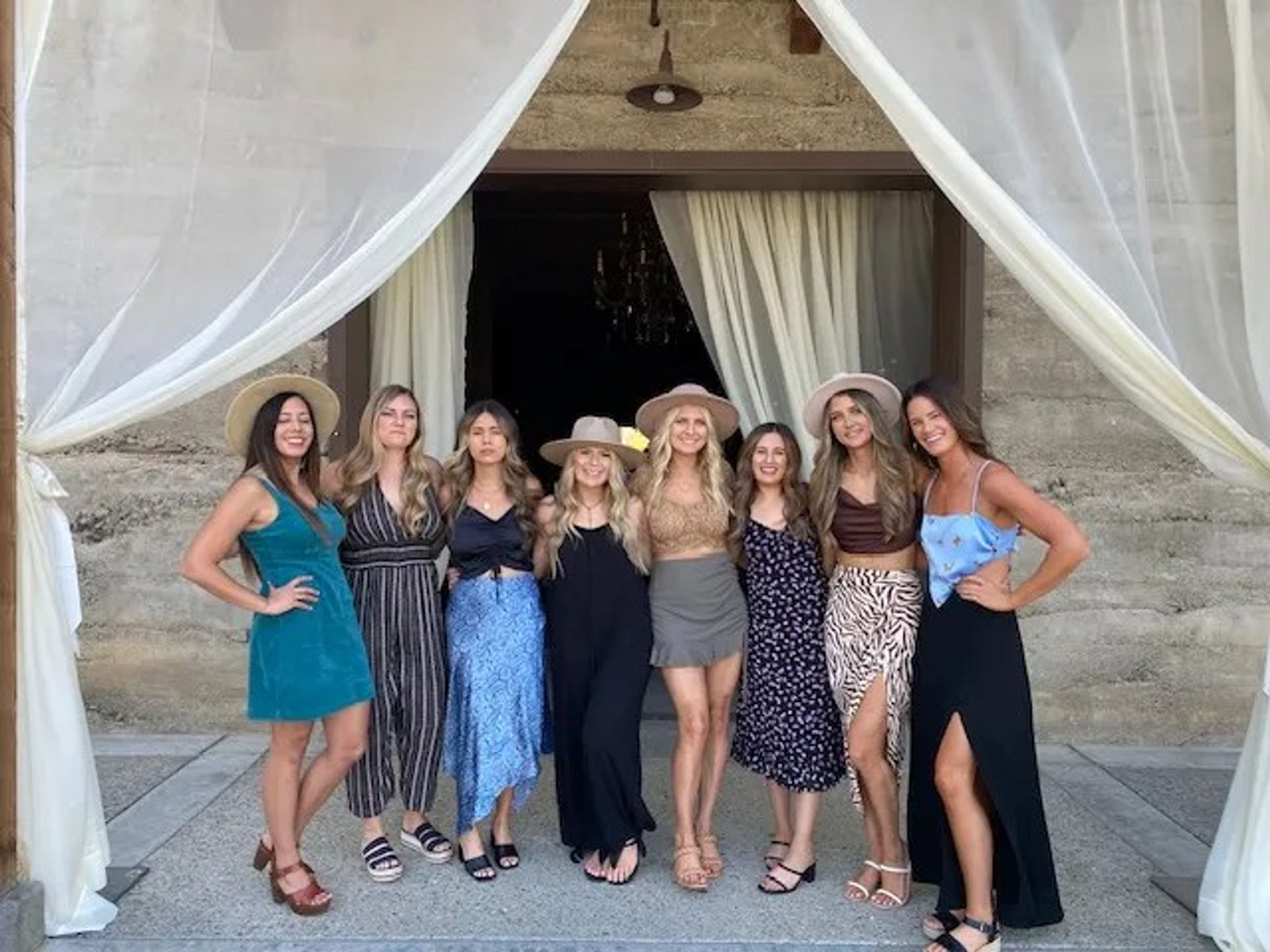 Smiling group portrait of eight women in summer dresses and sun hats posing under a stone archway framed by flowing sheer curtains