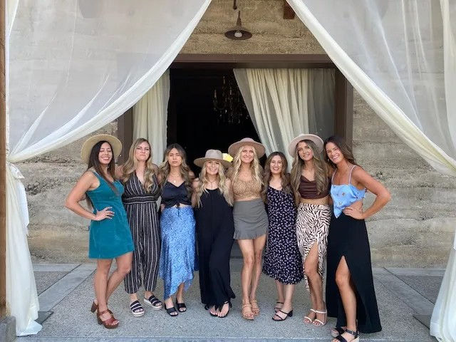 Smiling group portrait of eight women in summer dresses and sun hats posing under a stone archway framed by flowing sheer curtains