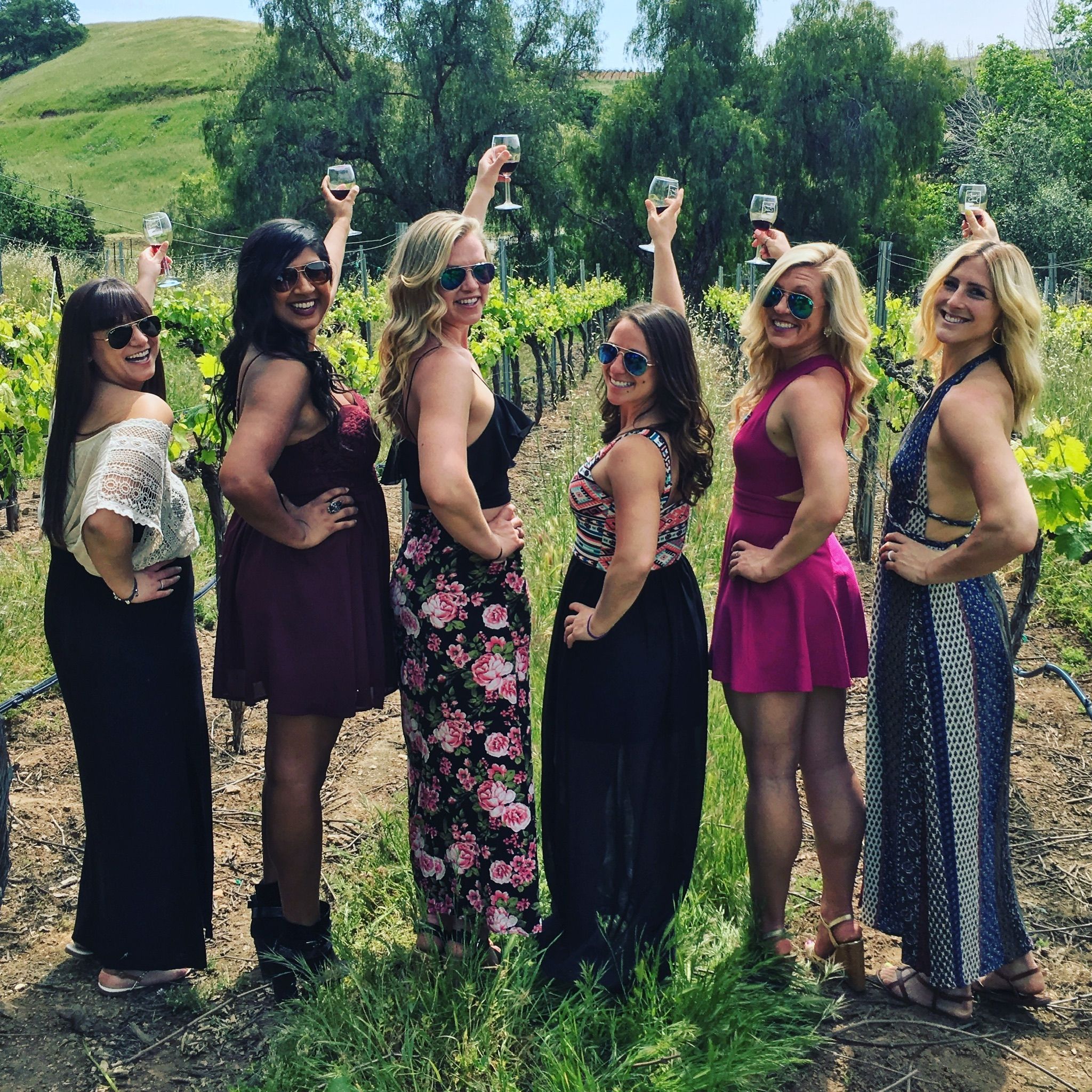 Six friends in sundresses raising wine glasses at a sunny vineyard wine-tasting amid green vines and rolling hills