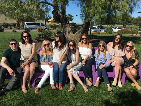 Cheerful group of friends in sunglasses and summer outfits sitting on purple lawn chairs under a large tree on a sunny grassy picnic lawn, smiling for a casual outdoor group photo.