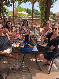 Women toasting with wine on a shaded vineyard patio during a sunny bachelorette wine-tasting, bride-to-be wearing sash and veil, plates of snacks and a bowl of chips on the table.