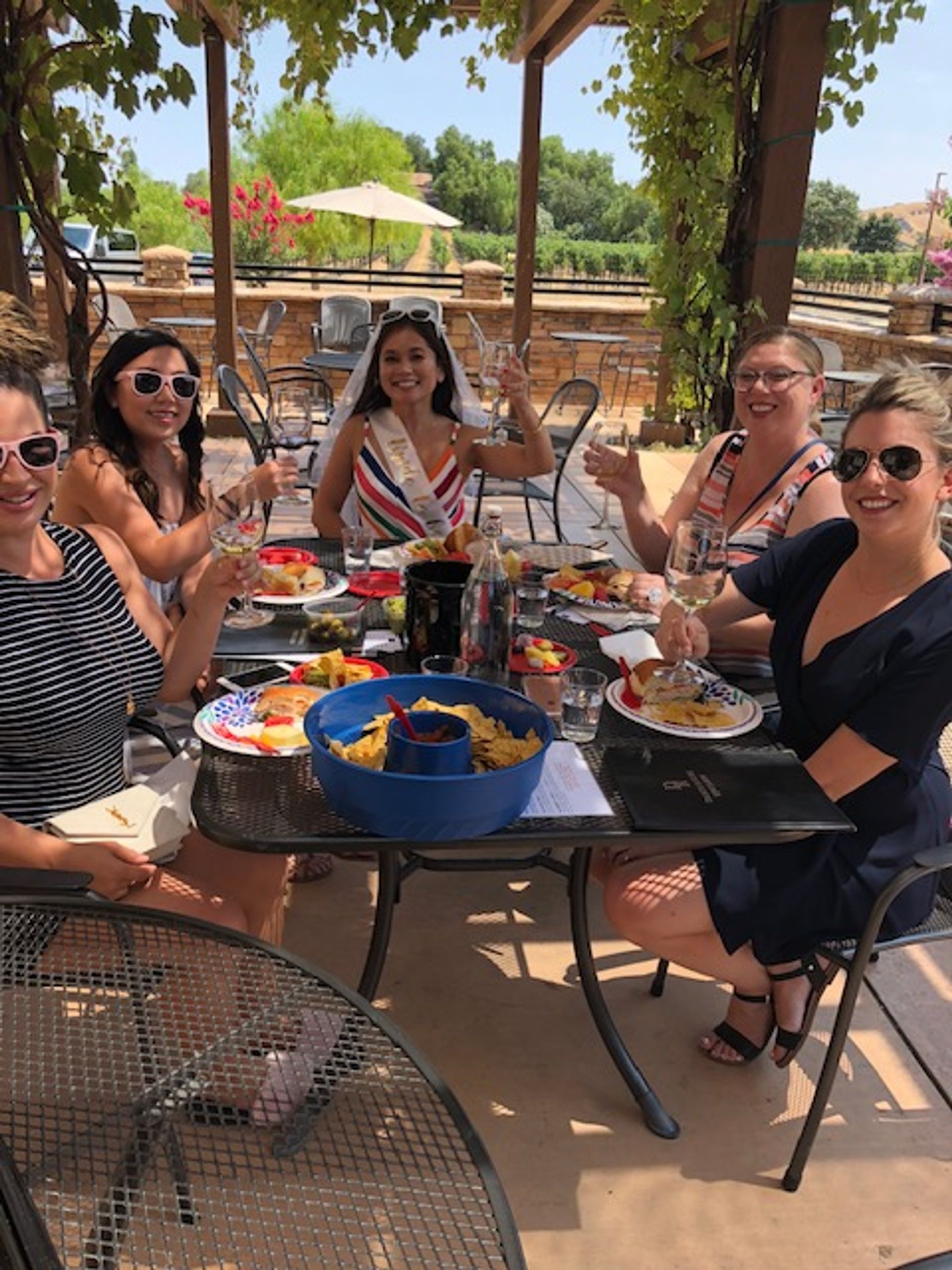 Group of women raising wine glasses at an outdoor vineyard patio brunch, bride-to-be wearing a sash and veil at center, plates of fruit, chips and summer dishes on a shaded pergola table.