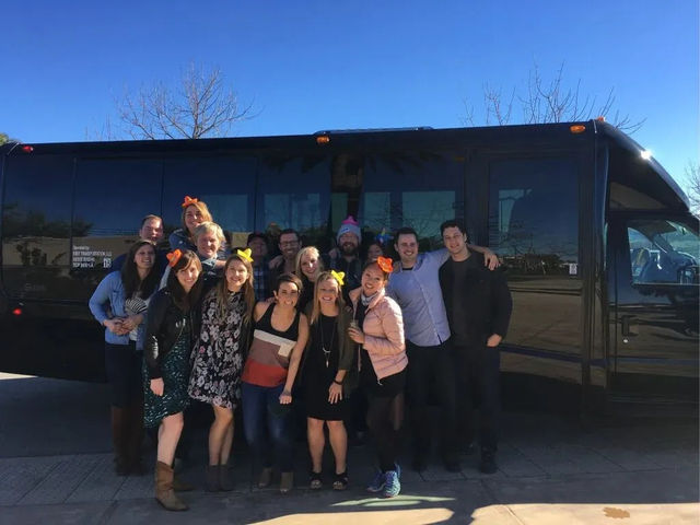 Group of about 15 adults smiling and posing in front of a black party/shuttle bus on a sunny day with a clear blue sky and bare trees, several wearing colorful bow headbands.