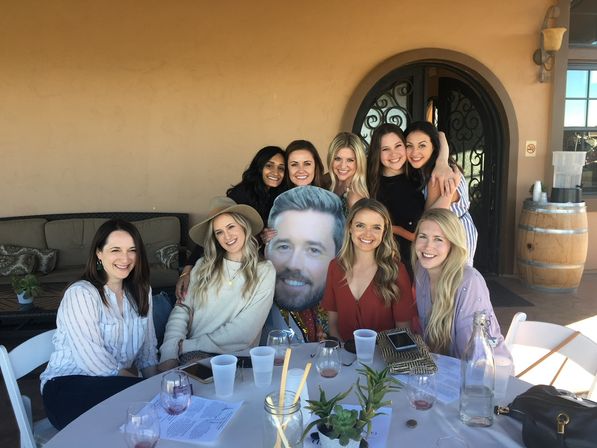 Smiling group of women at an outdoor winery patio wine tasting, seated around a round table with wine glasses, succulents, a wooden barrel, and a large humorous face cutout.