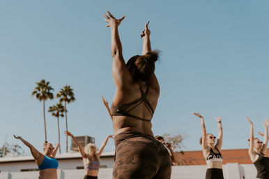 Outdoor group fitness workout in sunny Southern California — woman in camo leggings and strappy sports bra leads participants raising arms beneath palm trees.
