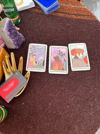 Three tarot cards (Nine of Swords, Seven of Swords, Page of Pentacles) laid on a brown woven tablecloth with an amethyst geode, a gold hand holding red cards, a blue card deck, and a green sparkling water can at an outdoor market stall.