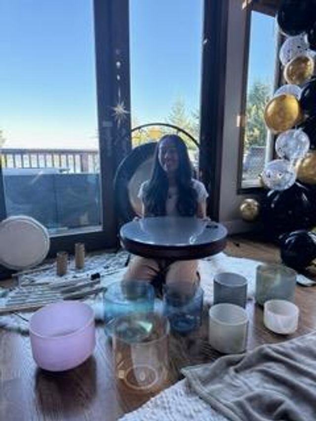 Person kneeling in an ocean-view living room playing a round handpan/steel drum, surrounded by crystal singing bowls and black-and-gold balloons near glass doors opening to a waterfront deck.