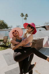 Two friends in bright sun hats and sunglasses laughing on a sunny backyard patio with palm trees, one hoisting the other in activewear and leggings near a hot tub.
