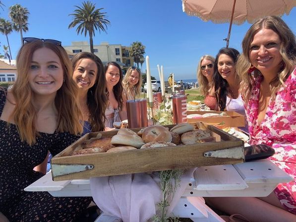 Smiling women at a sunny beachside brunch with a wooden tray of bagels on a picnic table, palm trees and ocean in the background.