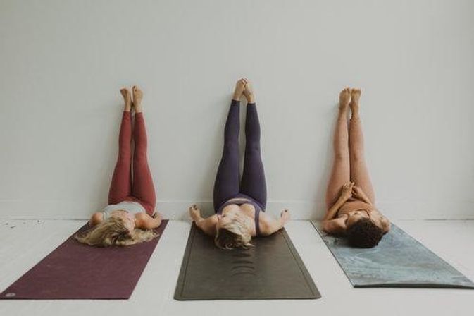 Three people in a bright, minimal yoga studio lying on mats with legs up the wall in restorative legs-up-the-wall pose, colorful leggings and mats on a white floor.