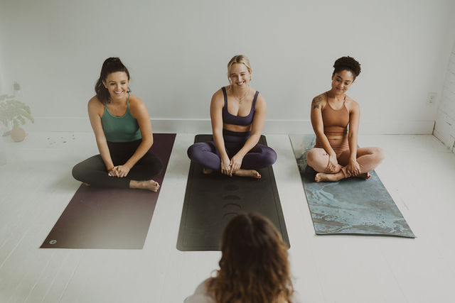 Three women in colorful activewear sitting cross-legged on yoga mats in a bright indoor yoga studio, smiling and relaxed during a friendly group yoga/wellness class.