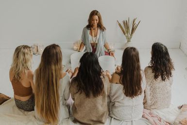 Women’s sound bath and guided meditation in a bright minimalist wellness studio — instructor plays white crystal singing bowls while five participants sit barefoot in a semicircle with candles and dried plants.