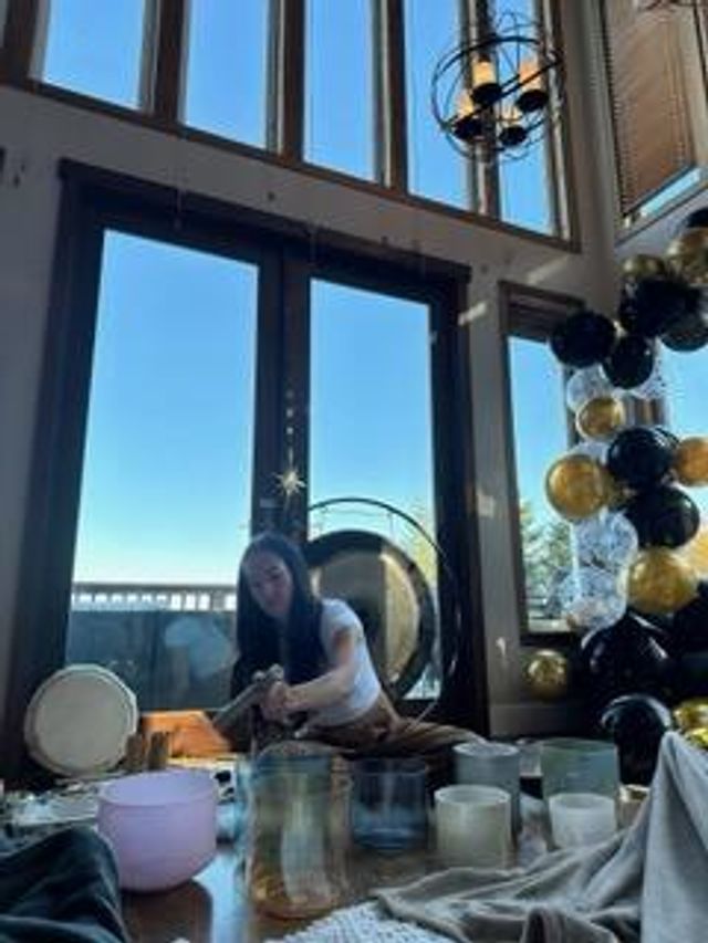 Sunlit high-ceiling studio with floor-to-ceiling windows; a person kneels arranging crystal singing bowls, a drum and a large gong next to a black-and-gold balloon arch — cozy sound‑healing setup.