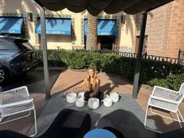 Person playing a white outdoor drum installation under a shaded pergola on a sunny open-air shopping center patio with modern chairs, blue-awning storefronts and palm trees