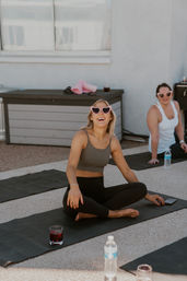 Two women laughing on yoga mats during a sunny rooftop outdoor yoga class, wearing pink heart-shaped sunglasses.