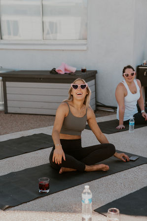 Two women laughing on yoga mats during a sunny rooftop outdoor yoga class, wearing pink heart-shaped sunglasses.