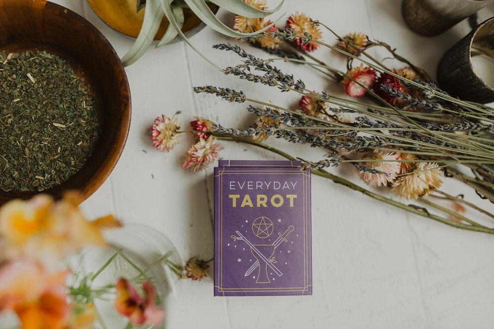 Boho flat lay of a purple tarot deck box on a white wooden table surrounded by dried lavender and strawflowers, a wooden bowl of dried herbs, and a glass vase with orange blooms.