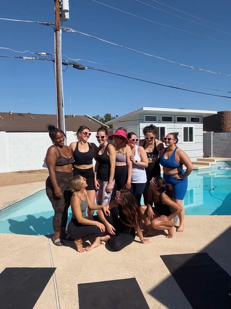 Group of women in activewear posing poolside at a suburban backyard pool on a sunny day, yoga mats on the deck — cheerful outdoor fitness gathering