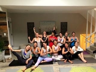 About a dozen people in a bright indoor yoga studio sitting on mats in two rows, smiling and striking playful flexed-arm poses during a group yoga class.