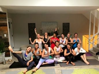 About a dozen people in a bright indoor yoga studio sitting on mats in two rows, smiling and striking playful flexed-arm poses during a group yoga class.