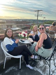 Three friends enjoying a casual rooftop hangout at sunset, seated around a small table with a vase of roses and wine glasses against an urban skyline.