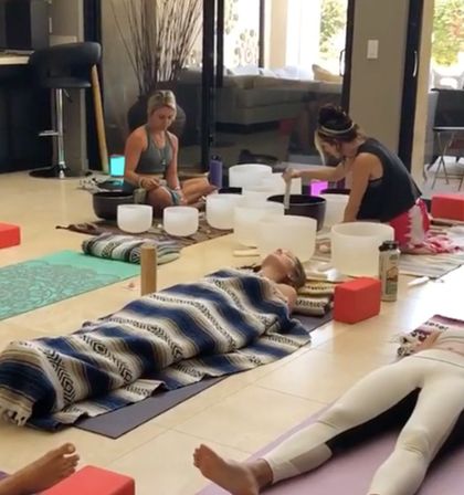 Participants reclined on yoga mats in an indoor wellness space—one wrapped in a striped blanket—while practitioners play crystal singing bowls during a relaxing sound bath meditation session.