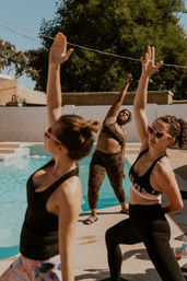 Three women doing a poolside outdoor yoga stretch at a sunny suburban backyard pool, instructor in camo leading while a participant wears heart-shaped sunglasses.