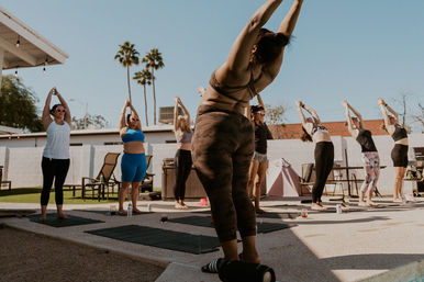 Sunny outdoor yoga class in a backyard courtyard—people on mats stretching upward beneath palm trees.