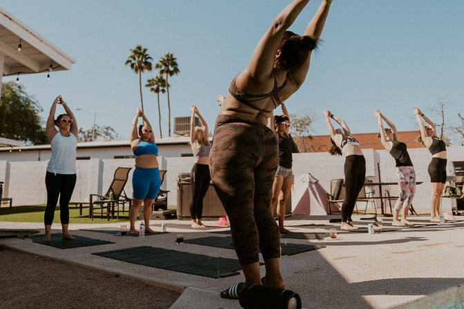 Sunny outdoor yoga class in a backyard courtyard—people on mats stretching upward beneath palm trees.