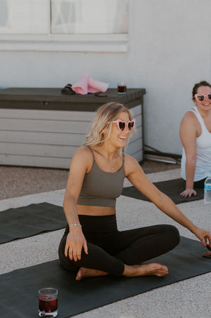 Smiling woman in pink heart-shaped sunglasses sitting cross-legged on a mat during a sunny outdoor rooftop yoga class, laughing with a drink and fellow participants nearby.