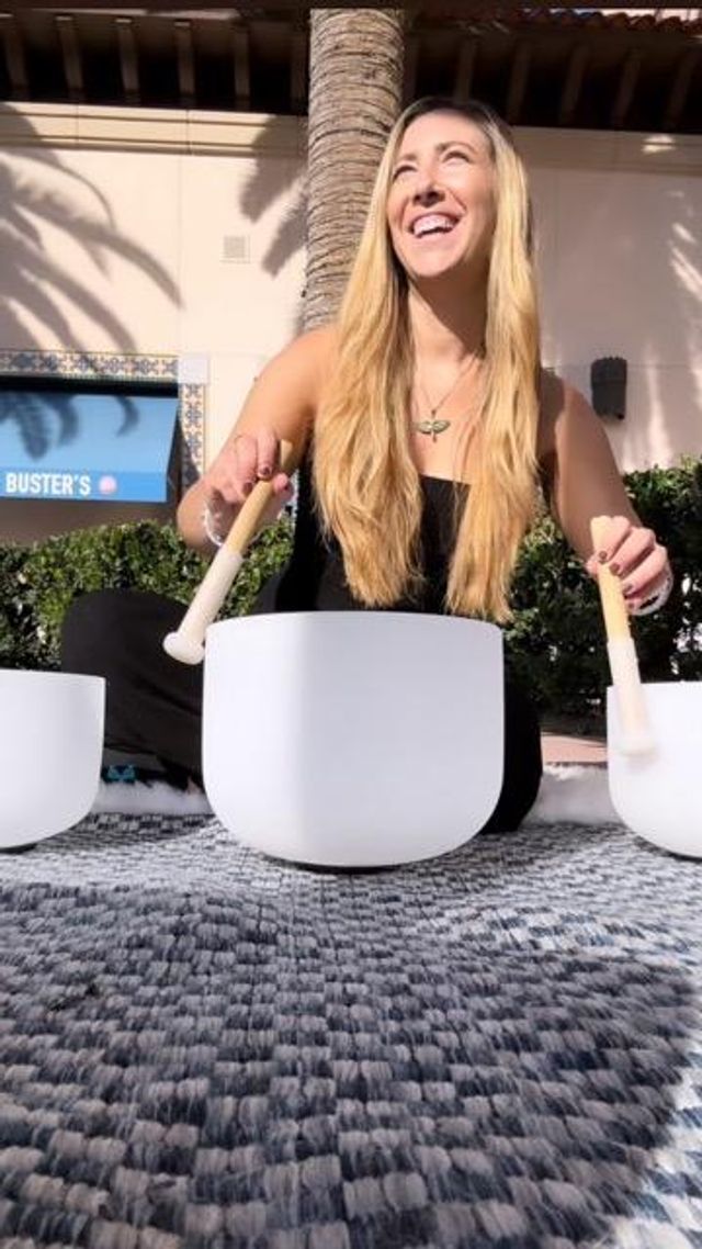 Smiling woman with long blonde hair playing white crystal singing bowls with mallets on a rug in a sunlit outdoor plaza under a palm tree