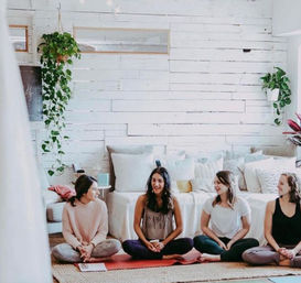 Four women sitting cross‑legged on yoga mats in a light‑filled boho studio lounge with white shiplap walls, hanging plants, cozy pillows and a woven rug — casual yoga/meditation circle.