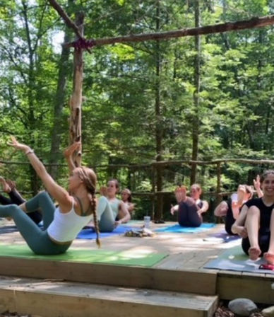 Sun-dappled outdoor group yoga class on a rustic wooden platform in a forest, participants on mats practicing boat pose beneath a leafy canopy