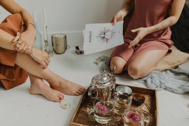 Two people seated on a white floor sharing a wooden tray with a silver teapot and glass cups of floral tea, one holding a booklet.