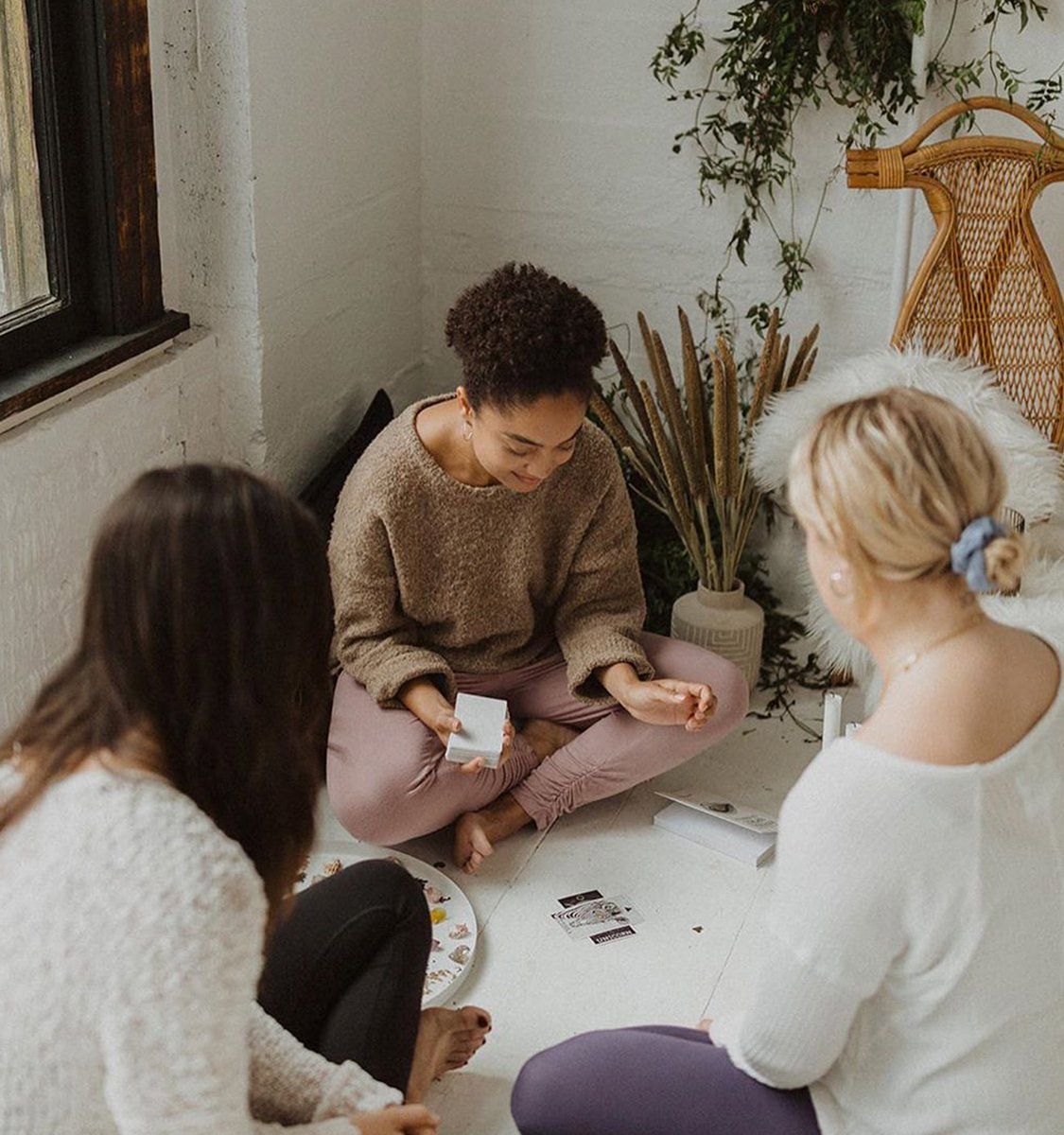 Three women seated cross-legged on a sunlit boho floor, sharing and reading cards amid plants and a rattan chair in a cozy studio-style living room