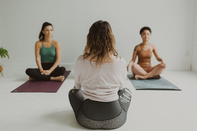 Yoga instructor with back to camera leading two students seated cross-legged on mats in a bright, minimalist studio