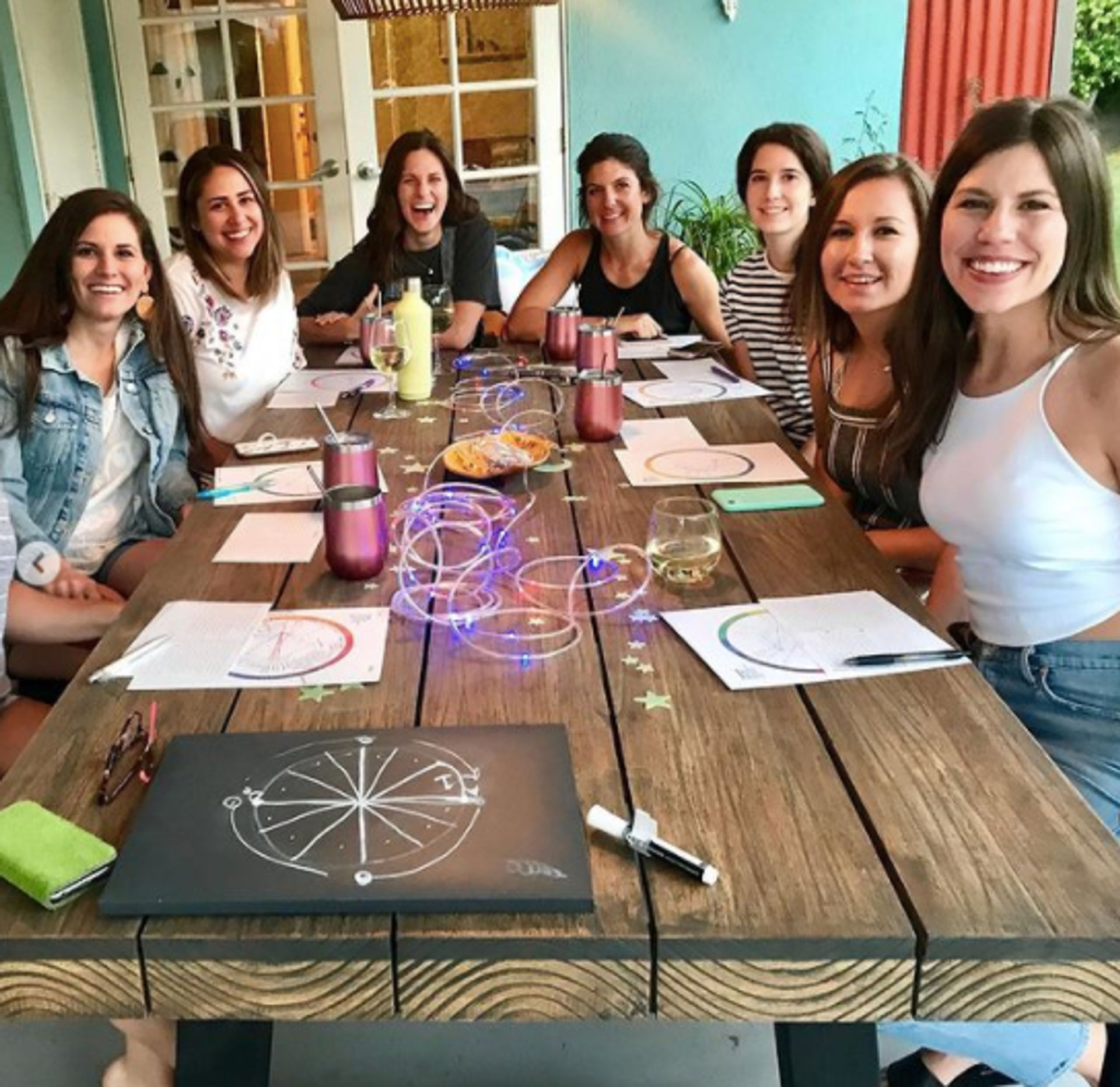 Seven friends smiling around a wooden outdoor patio table having a fun craft/vision-board night with papers, markers, drinks, string lights and snacks